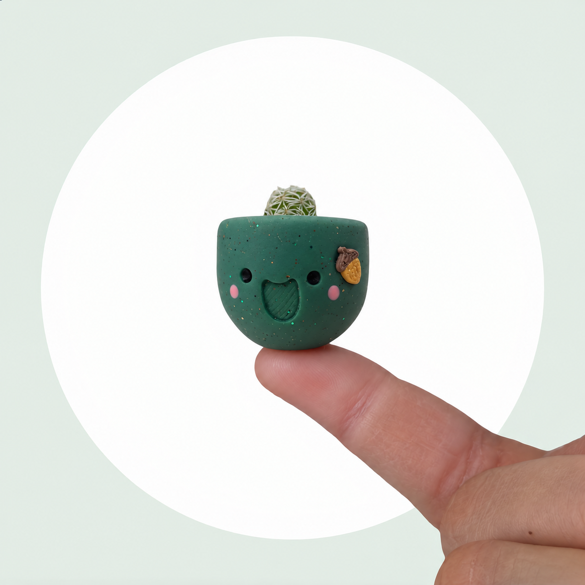 Acorny live mini cactus in a handmade sage green pot with rosy cheeks and a tiny acorn, balanced on a fingertip