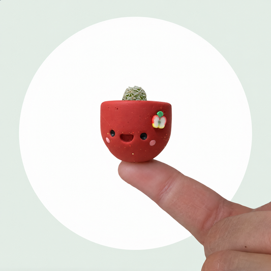 Apple Cider live mini cactus in a handmade red pot with rosy cheeks and a little apple slice, balanced on a fingertip