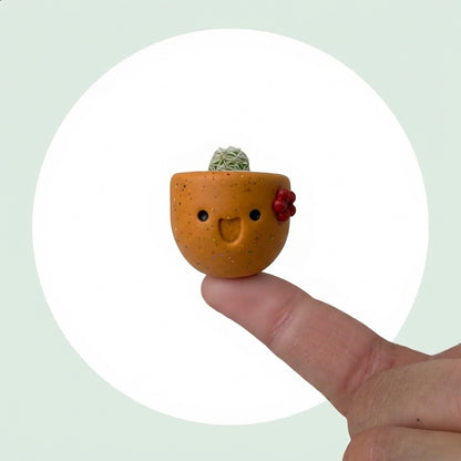 Pumpkin Spice live mini cactus in a handmade pumpkin orange pot with a maroon flower, balanced on a fingertip